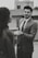 A man in a suit gently holding the hand of another person, possibly engaging in a moment of connection or conversation. The setting appears to be urban, with blurred buildings in the background. The image is in black and white, adding a classic or timeless feel.