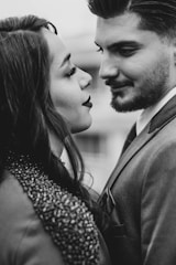 A couple facing each other closely, suggesting intimacy and affection. The woman has long hair and is wearing a textured garment, while the man has a neatly trimmed beard and is dressed in a suit. The image is in black and white, emphasizing contrast and depth.