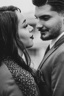 A couple facing each other closely, suggesting intimacy and affection. The woman has long hair and is wearing a textured garment, while the man has a neatly trimmed beard and is dressed in a suit. The image is in black and white, emphasizing contrast and depth.