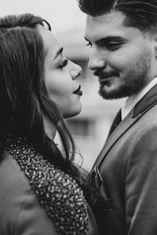 A couple facing each other closely, suggesting intimacy and affection. The woman has long hair and is wearing a textured garment, while the man has a neatly trimmed beard and is dressed in a suit. The image is in black and white, emphasizing contrast and depth.