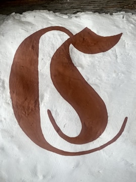 A large, stylized brown letter resembling a 'C' is painted on a textured white surface. The letter has a calligraphic, artistic design with a curving tail extending from the top.