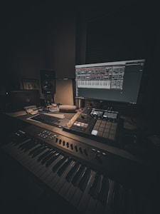 A dimly lit music production setup featuring a keyboard, a monitor displaying music software, a mixer, and various control pads. The room has a cozy atmosphere with a focus on digital music creation. Cables and a laptop are also visible, suggesting a workspace dedicated to audio work.