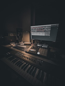 A dimly lit music production setup featuring a keyboard, a monitor displaying music software, a mixer, and various control pads. The room has a cozy atmosphere with a focus on digital music creation. Cables and a laptop are also visible, suggesting a workspace dedicated to audio work.