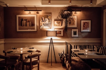 A cozy, dimly lit dining area featuring a mix of round and rectangular tables set with napkins and glasses. The walls are adorned with framed artwork, including portraits and abstract pieces. A minimalist floor lamp with a cylindrical shade stands beside the seating area. The overall ambiance is warm and intimate, enhanced by the soft glow of candles on the tables.