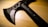 A black tactical tomahawk sits on a wooden surface. The tomahawk features a sharp blade with a serrated edge and several circular cutouts for weight reduction. The handle is made of a durable-looking material, with visible rivet-like fasteners.