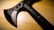 A black tactical tomahawk sits on a wooden surface. The tomahawk features a sharp blade with a serrated edge and several circular cutouts for weight reduction. The handle is made of a durable-looking material, with visible rivet-like fasteners.