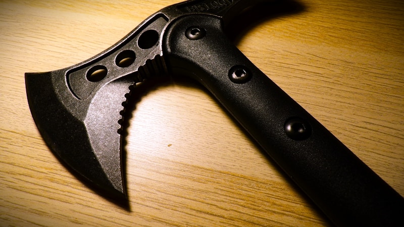 A black tactical tomahawk sits on a wooden surface. The tomahawk features a sharp blade with a serrated edge and several circular cutouts for weight reduction. The handle is made of a durable-looking material, with visible rivet-like fasteners.