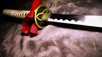 A close-up view of a traditional Japanese katana with a decorative red and gold handguard, featuring intricate patterns. The sword's blade is sharp and polished, reflecting light subtly. The handle is wrapped in a beige cord, adding texture to the otherwise smooth appearance of the sword.
