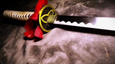 A close-up view of a traditional Japanese katana with a decorative red and gold handguard, featuring intricate patterns. The sword's blade is sharp and polished, reflecting light subtly. The handle is wrapped in a beige cord, adding texture to the otherwise smooth appearance of the sword.