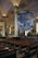 Inside a large, ornate church with high arched ceilings, there's an art installation featuring a giant globe suspended in the middle, surrounded by chandeliers. The church interior includes rows of wooden pews, large supporting columns, and intricate wrought iron details near the altar area. Stained glass windows allow natural light to filter through.
