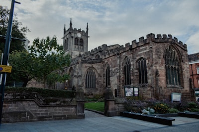 A historic stone church with Gothic architectural features, including arched windows and a prominent clock tower, surrounded by trees and a well-maintained garden. The setting is urban, indicated by nearby buildings and a pedestrian zone sign.