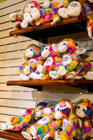 Colorful stuffed monkeys with tie-dye patterns are displayed on wooden shelves against a light-colored wall. Each monkey has white hands and feet and a friendly face with embroidered features. Some have tags attached.
