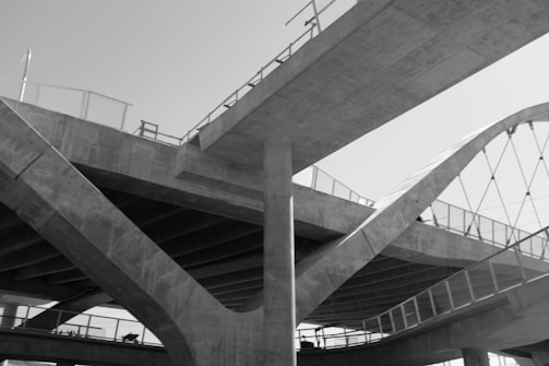 A black and white photograph of a modern architectural structure featuring multiple intersecting bridges and walkways, supported by large concrete pillars. The design includes curved and angular elements with railings and a network of cables.