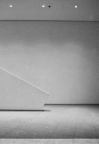 A minimalist, monochromatic interior space featuring a clean, angular design with a diagonal wall or partition. The scene is well-lit by ceiling spotlights, and the floor is covered with a textured surface.