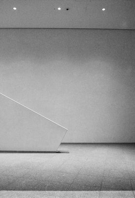 A minimalist, monochromatic interior space featuring a clean, angular design with a diagonal wall or partition. The scene is well-lit by ceiling spotlights, and the floor is covered with a textured surface.