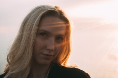 A woman with long blonde hair is captured against a bright, softly blurred background. Sunlight creates a warm glow around her and casts rays across her face, adding a serene and ethereal quality to the image.