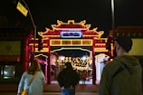 A vibrant Chinatown gate is lit up with bright neon lights at night. Several people are walking towards the gate, which is adorned with intricate traditional architectural designs. Colorful lanterns hang in the background, enhancing the festive atmosphere.