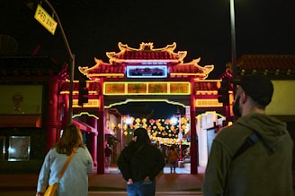 A vibrant Chinatown gate is lit up with bright neon lights at night. Several people are walking towards the gate, which is adorned with intricate traditional architectural designs. Colorful lanterns hang in the background, enhancing the festive atmosphere.
