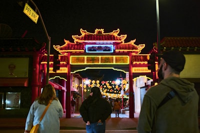 A vibrant Chinatown gate is lit up with bright neon lights at night. Several people are walking towards the gate, which is adorned with intricate traditional architectural designs. Colorful lanterns hang in the background, enhancing the festive atmosphere.