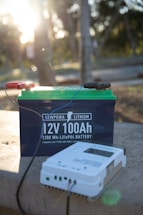 A 12V 100Ah lithium battery with visible branding and a connected device on a stone surface in an outdoor setting. The background is softly blurred, showing sunlight filtering through trees, creating a serene environment.
