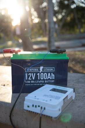 A 12V 100Ah lithium battery with visible branding and a connected device on a stone surface in an outdoor setting. The background is softly blurred, showing sunlight filtering through trees, creating a serene environment.