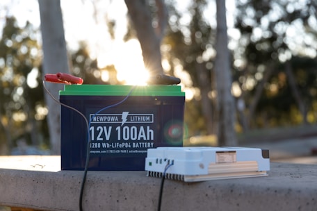A lithium battery labeled 'Newpowa 12V 100Ah' sits on a concrete surface outdoors. It is connected by cables to a separate white electrical device. The background features blurred trees with sunlight filtering through, creating a warm, natural ambiance.