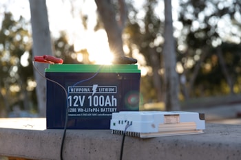 A lithium battery labeled 'Newpowa 12V 100Ah' sits on a concrete surface outdoors. It is connected by cables to a separate white electrical device. The background features blurred trees with sunlight filtering through, creating a warm, natural ambiance.