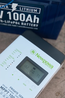 A Newpowa MPPT 100 solar charge controller is placed near a 100Ah lithium battery. The controller's display shows readings of 13.2 volts, 0.0 amps, and 77%. The battery, marked as LiFePO4, is dark blue with white text highlighting its specifications.