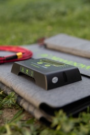 A device with a black interface, displaying green digital numbers and symbols, rests on a folded grey fabric surface. To its left, a coil of red and black cables is partially visible, and the background features green grass, indicating an outdoor setting.
