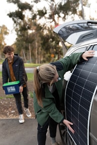 A person is placing a large solar panel against the side of a car with an open trunk, while another person stands nearby holding a plastic container with handles. Trees and greenery are visible in the blurred background.