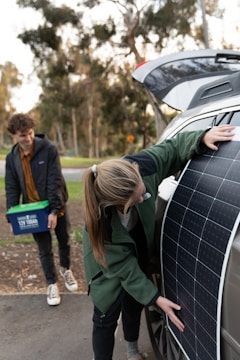 A person is placing a large solar panel against the side of a car with an open trunk, while another person stands nearby holding a plastic container with handles. Trees and greenery are visible in the blurred background.
