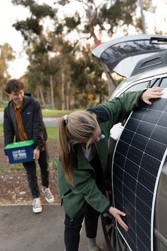 A person is placing a large solar panel against the side of a car with an open trunk, while another person stands nearby holding a plastic container with handles. Trees and greenery are visible in the blurred background.