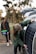 A person is placing a large solar panel against the side of a car with an open trunk, while another person stands nearby holding a plastic container with handles. Trees and greenery are visible in the blurred background.