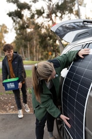 A person is placing a large solar panel against the side of a car with an open trunk, while another person stands nearby holding a plastic container with handles. Trees and greenery are visible in the blurred background.