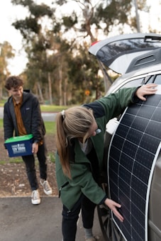 A person is placing a large solar panel against the side of a car with an open trunk, while another person stands nearby holding a plastic container with handles. Trees and greenery are visible in the blurred background.