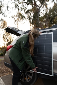 A person with long hair, wearing a green jacket, is adjusting or holding a solar panel. The panel is leaning against a silver car with an open trunk. The surroundings include trees and a dimly lit, outdoor setting, likely during sunset.