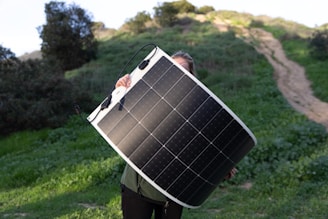 A person holds a large flexible solar panel in front of their body, standing outdoors on lush green grass. The background features a hill with trees and a dirt path winding up the slope.
