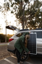 A person is setting up a solar panel against the side of a vehicle. They are wearing a green jacket and have long hair tied back. The car is parked outdoors with the hatchback open, and there are trees and a clear sky in the background.
