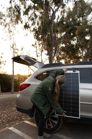 A person is setting up a solar panel against the side of a vehicle. They are wearing a green jacket and have long hair tied back. The car is parked outdoors with the hatchback open, and there are trees and a clear sky in the background.