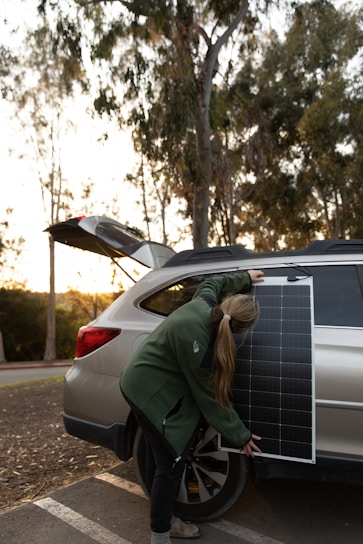 A person is setting up a solar panel against the side of a vehicle. They are wearing a green jacket and have long hair tied back. The car is parked outdoors with the hatchback open, and there are trees and a clear sky in the background.