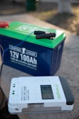 A large blue and green lithium battery with clamps attached is positioned on a flat surface next to a Newpowa solar charge controller. The controller is white with a digital display and various buttons and indicators.