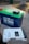 A large blue and green lithium battery with clamps attached is positioned on a flat surface next to a Newpowa solar charge controller. The controller is white with a digital display and various buttons and indicators.