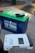 A large blue and green lithium battery with clamps attached is positioned on a flat surface next to a Newpowa solar charge controller. The controller is white with a digital display and various buttons and indicators.