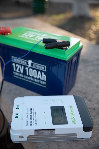 A large blue and green lithium battery with clamps attached is positioned on a flat surface next to a Newpowa solar charge controller. The controller is white with a digital display and various buttons and indicators.