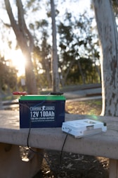 A lithium battery labeled '12V 100Ah' rests on a concrete bench in an outdoor setting with trees and sunlight visible. Wires are attached to the battery, and beside it is a white device, possibly a charger.