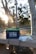 A lithium battery labeled '12V 100Ah' rests on a concrete bench in an outdoor setting with trees and sunlight visible. Wires are attached to the battery, and beside it is a white device, possibly a charger.