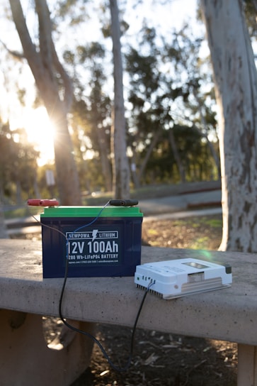 A lithium battery labeled '12V 100Ah' rests on a concrete bench in an outdoor setting with trees and sunlight visible. Wires are attached to the battery, and beside it is a white device, possibly a charger.