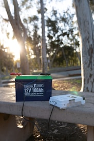 A lithium battery labeled '12V 100Ah' rests on a concrete bench in an outdoor setting with trees and sunlight visible. Wires are attached to the battery, and beside it is a white device, possibly a charger.