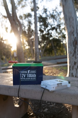 A lithium battery labeled '12V 100Ah' rests on a concrete bench in an outdoor setting with trees and sunlight visible. Wires are attached to the battery, and beside it is a white device, possibly a charger.