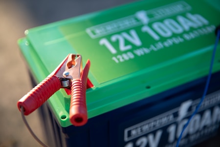 A close-up view of a car battery with a prominent red jumper cable clamp attached to it. The battery has a green top with white text displaying '12V 100Ah'. A cable is seen extending from the clamp, suggesting it is in use.